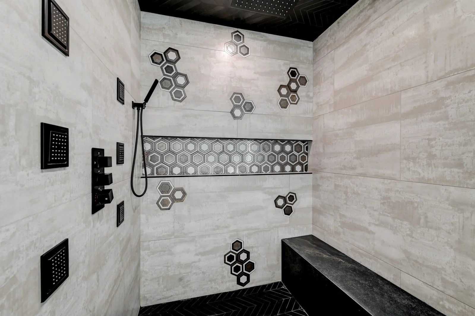 Shower with gray porcelain tile walls, hexagonal accent band, black fixtures, and stone bench