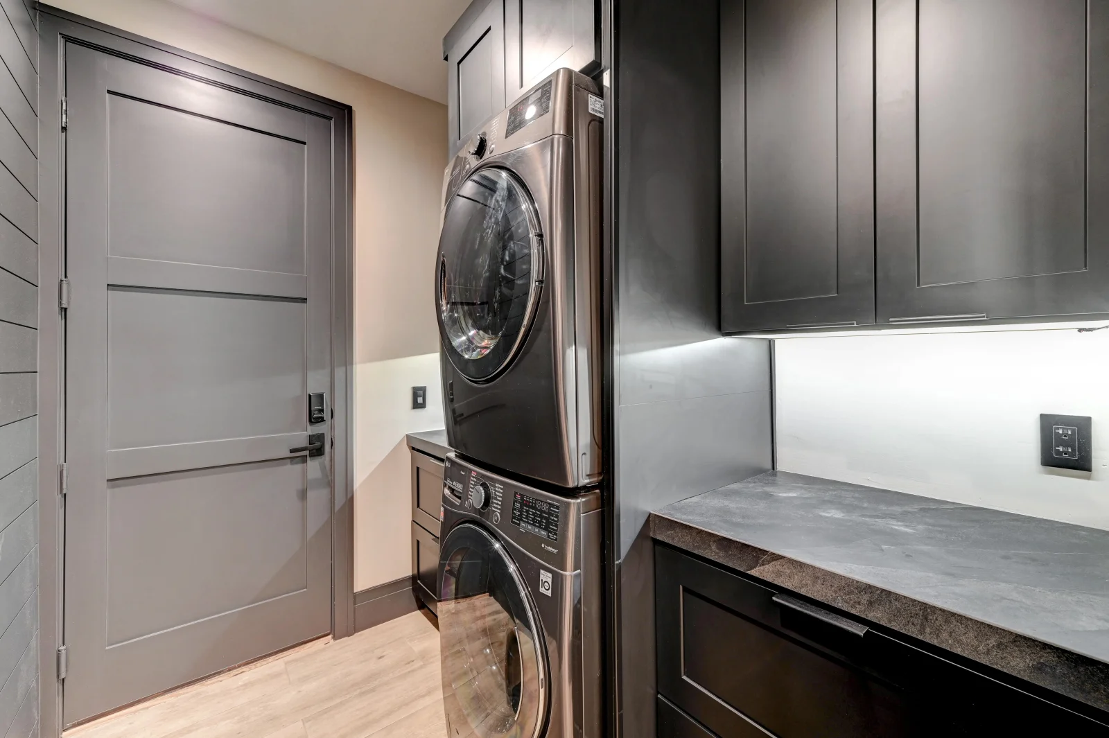 Modern laundry room with dark cabinetry, quartz countertop, stacked washer-dryer, and white subway tile backsplash