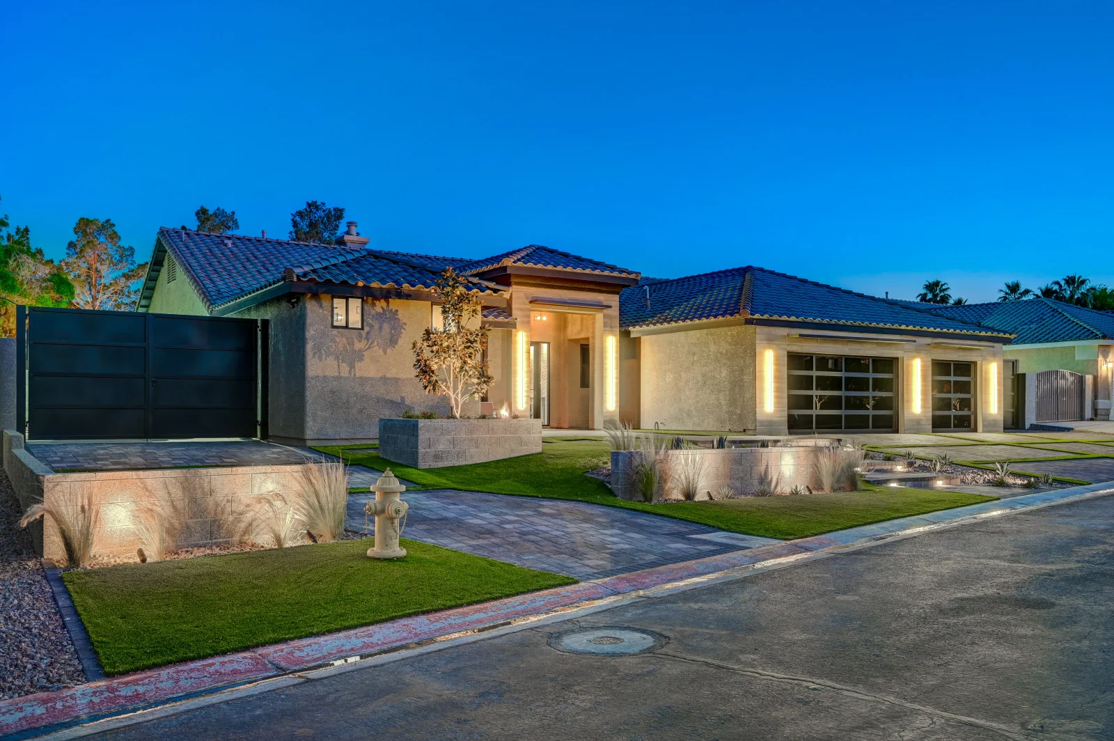 Modern home with dark metal tile roof, stucco walls, paver driveway, and landscape lighting