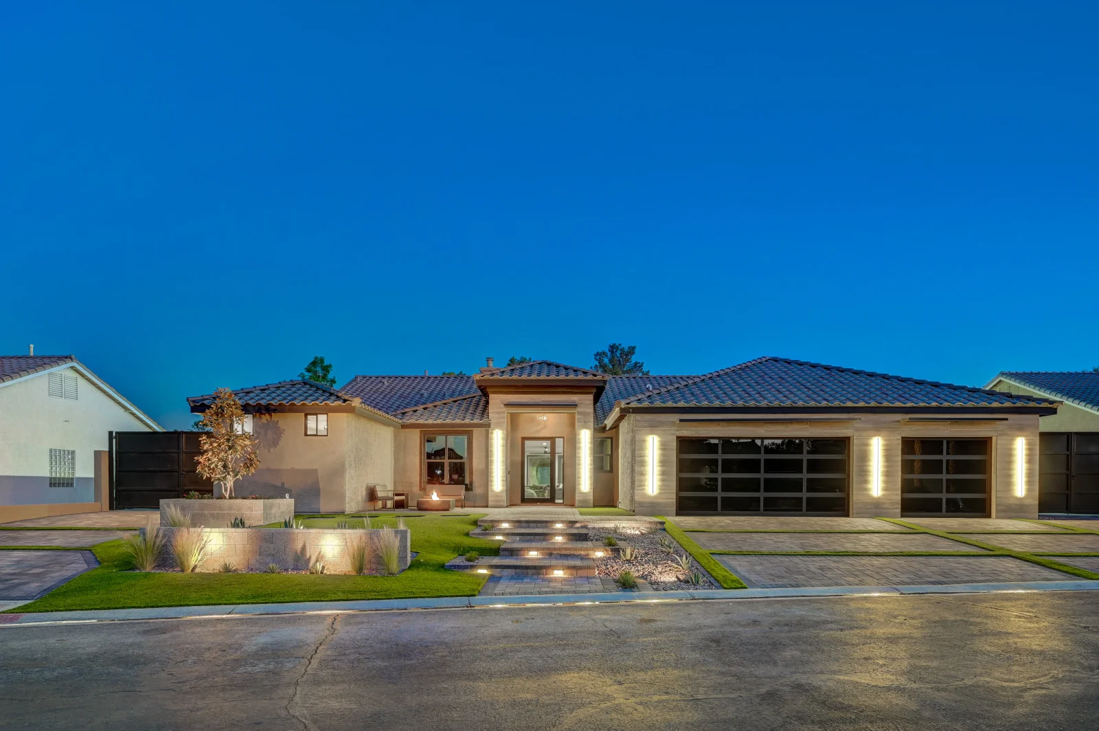 Contemporary stucco home with dark tile roof, glass garage doors, lit stone pathway, and modern landscaping