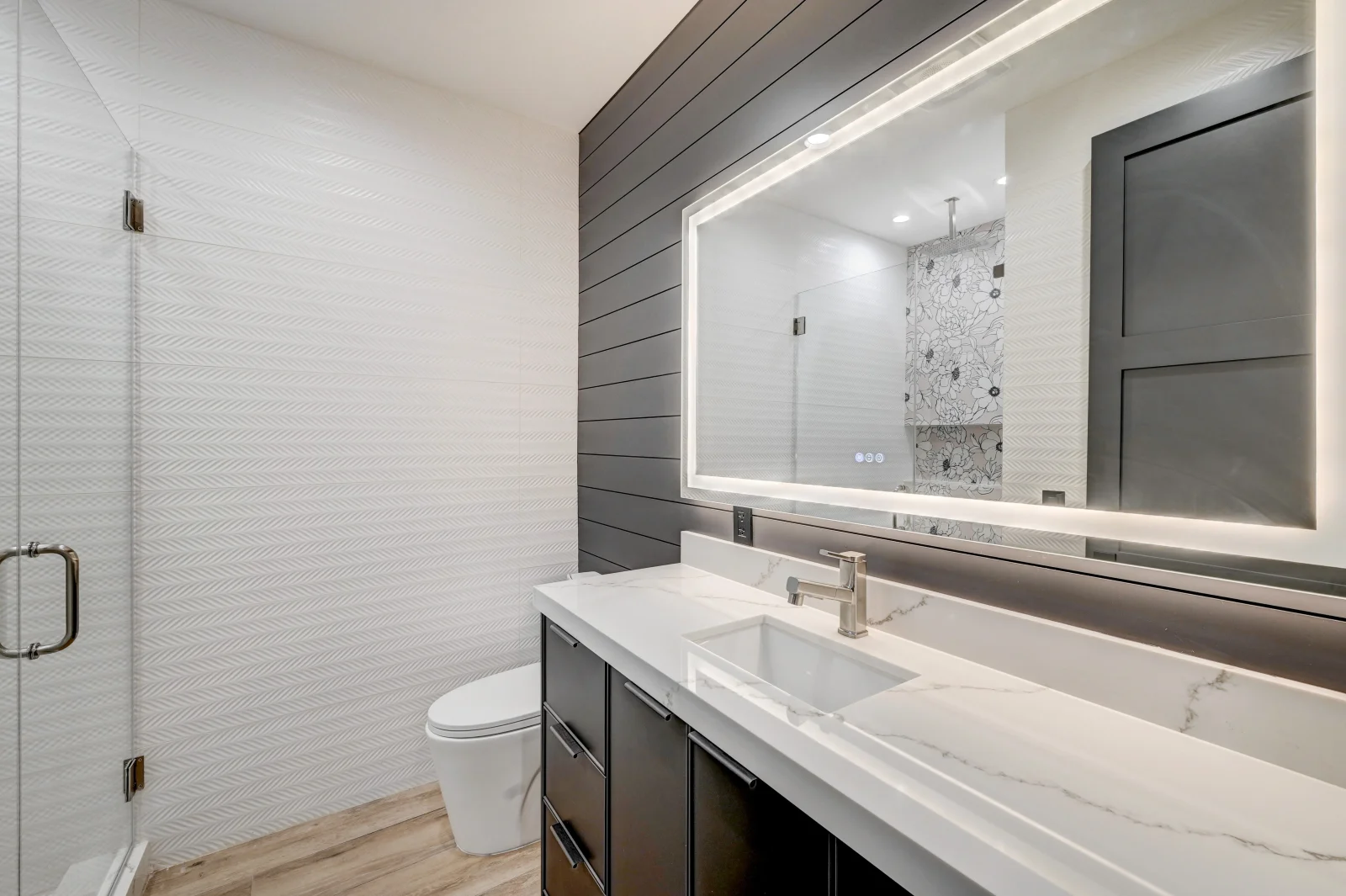 Modern bathroom with herringbone tile, marble countertop, frameless glass shower, and illuminated mirror